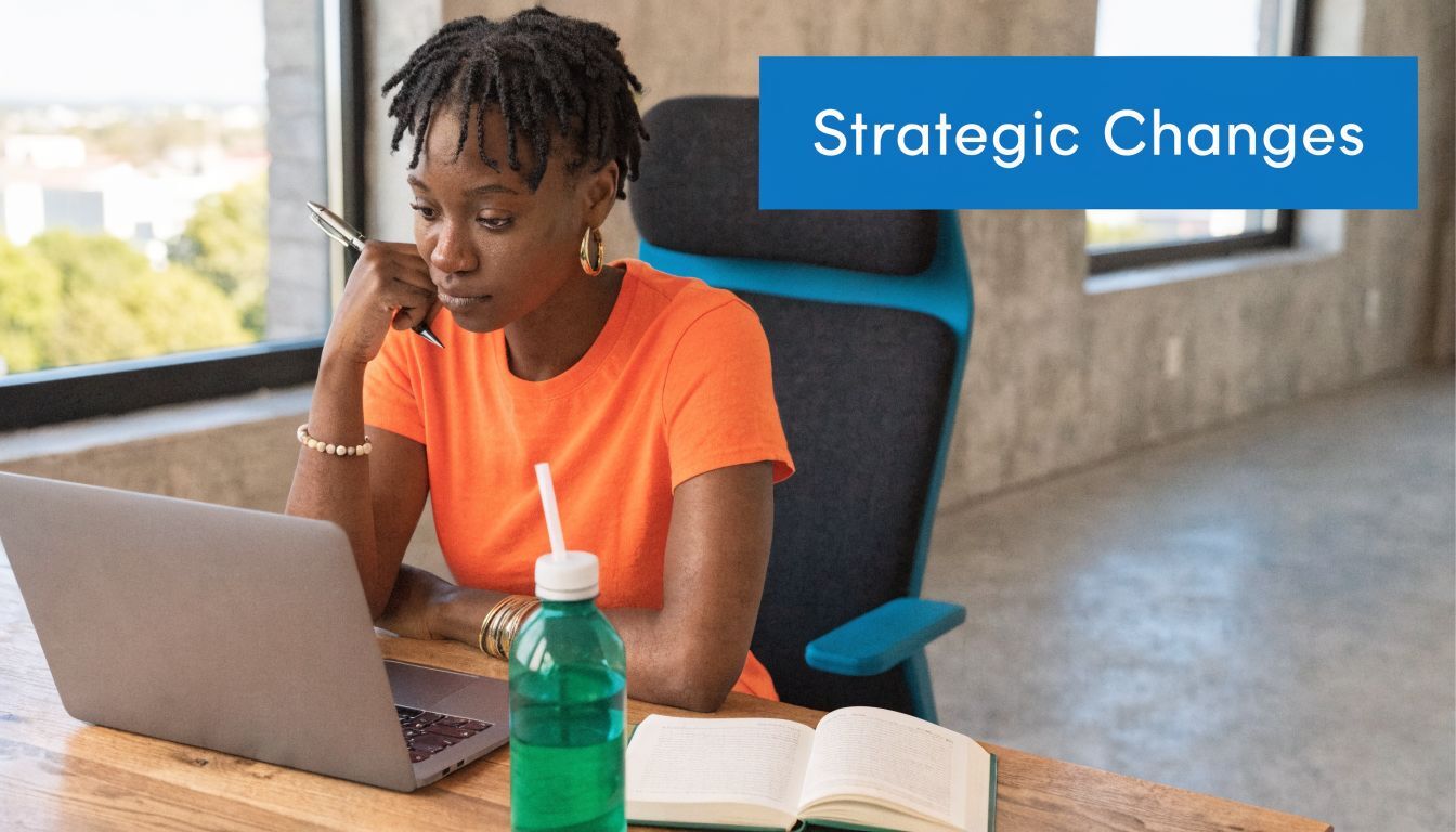 A young professional woman in an orange shirt working on her laptop with a book and water bottle.